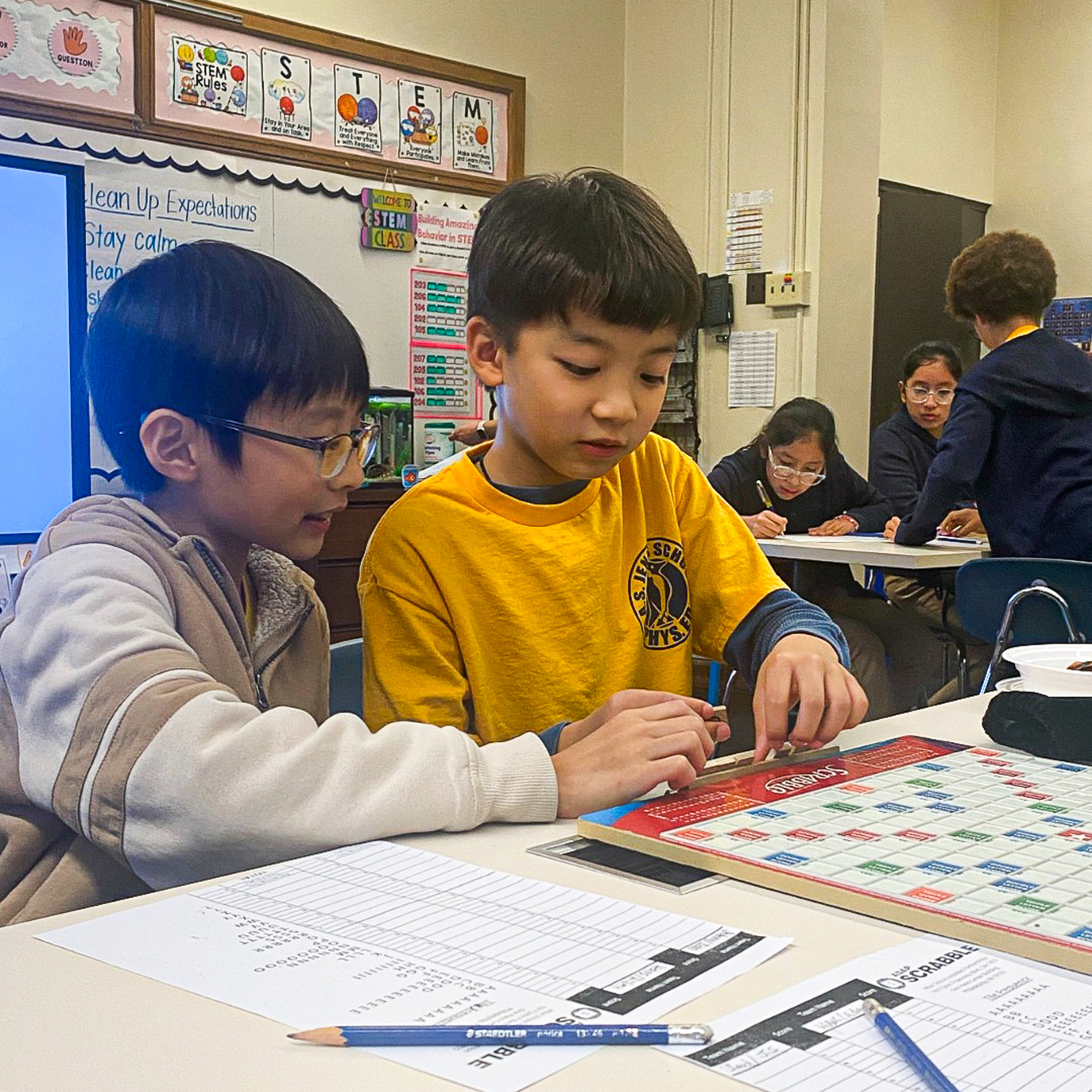 Students Collaborate During STEM Activity at Abram Jenks School Two students at Abram Jenks School work together on a Scrabble board during a STEM classroom activity while classmates collaborate in the background.