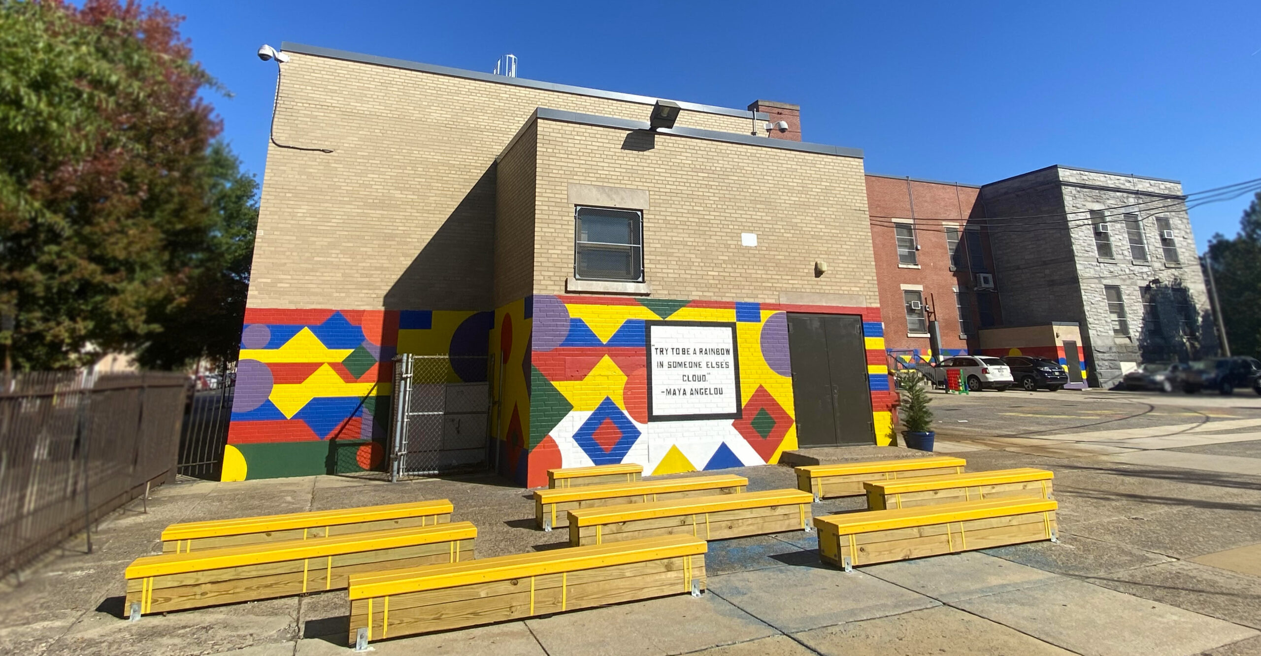 Abram Jenks School Building and Community Mural Exterior view of Abram Jenks School featuring a colorful geometric mural on the building, yellow seating in the schoolyard, and a quote displayed on the wall under a clear blue sky.